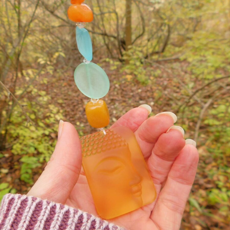 A hand holding a suncatcher with a Buddha-shaped amber-colored glass pendant. The suncatcher features vibrant beads in orange, blue, and aqua hues. The background shows a peaceful, autumn forest with golden leaves and soft natural lighting.</p>
<p>