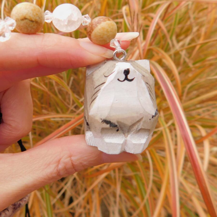 A hand holding a car charm featuring a hand-carved wooden cat pendant with a whimsical, painted face. The charm includes natural stone beads in earthy tones and clear crystal accents. The background showcases tall ornamental grasses with warm autumn colors.